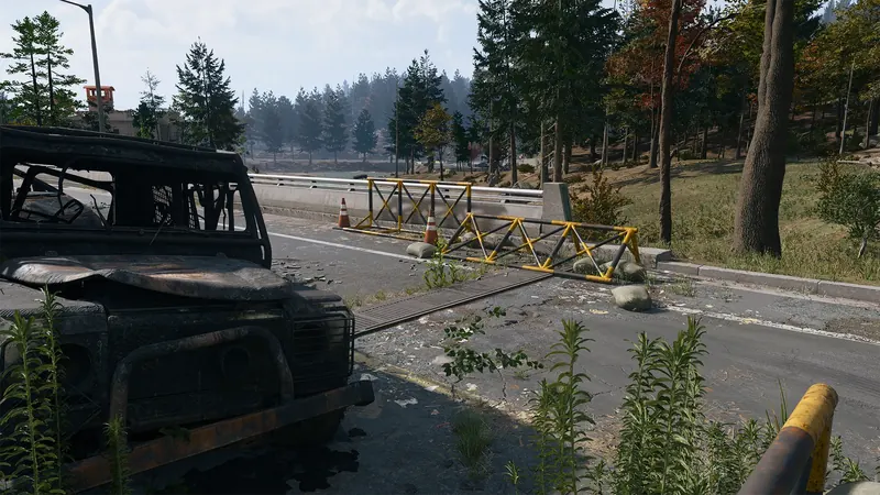 Burned-out military vehicle sits beside yellow road barricades near a wooded riverside.