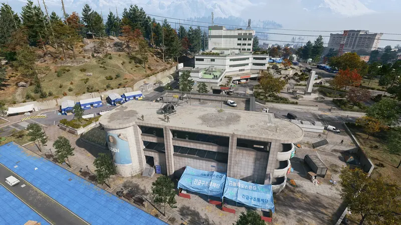 Aerial view of a hospital complex with a helicopter on its rooftop, emergency tents, and wooded hills nearby.